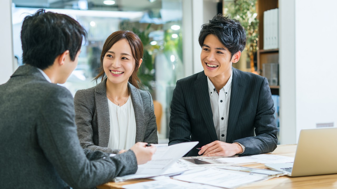 Modern ofis ortamında, Japon emlakçı ve genç kiracı çifti kira sözleşmesi detaylarını anlatırken, güven ve şeffaflık vurgusu.