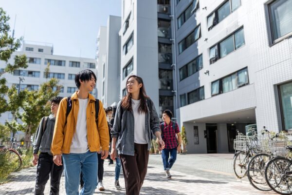 Osaka'daki modern öğrenci yurdu önünde farklı kökenlerden gençler, bisiklet ve sırt çantasıyla yürüyüş yaparken, şehir manzarasıyla birlikte.
