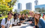 Tokyo Üniversitesi kampüsünde, açık havada sırt çantalı ve dizüstü bilgisayarlı uluslararası öğrenciler birlikte gülümseyerek ders çalışıyor.