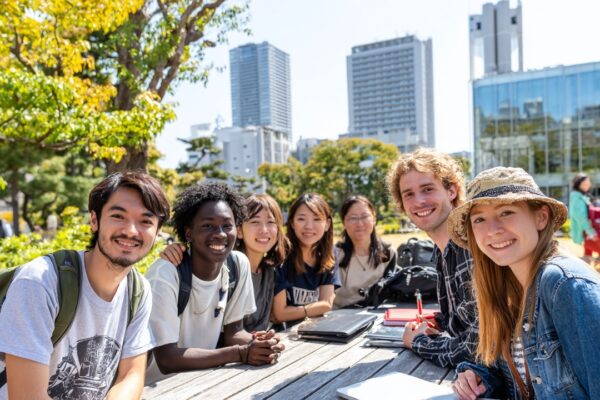 Tokyo Üniversitesi kampüsünde, açık havada sırt çantalı ve dizüstü bilgisayarlı uluslararası öğrenciler birlikte gülümseyerek ders çalışıyor.