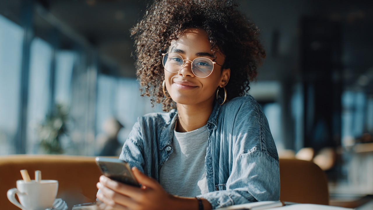 Genç uluslararası öğrenci, finansal farkındalık ve akıllı bütçeleme için cep telefonu uygulamasıyla giderleri takip ederken modern kafe veya öğrenci salonunda oturuyor.